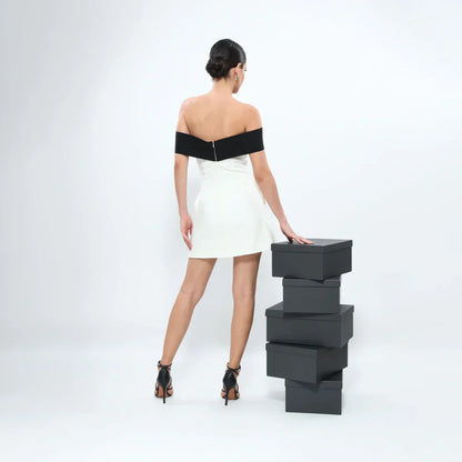 Back view of a model wearing a classic black and white off-shoulder dress, standing next to a stack of black boxes.