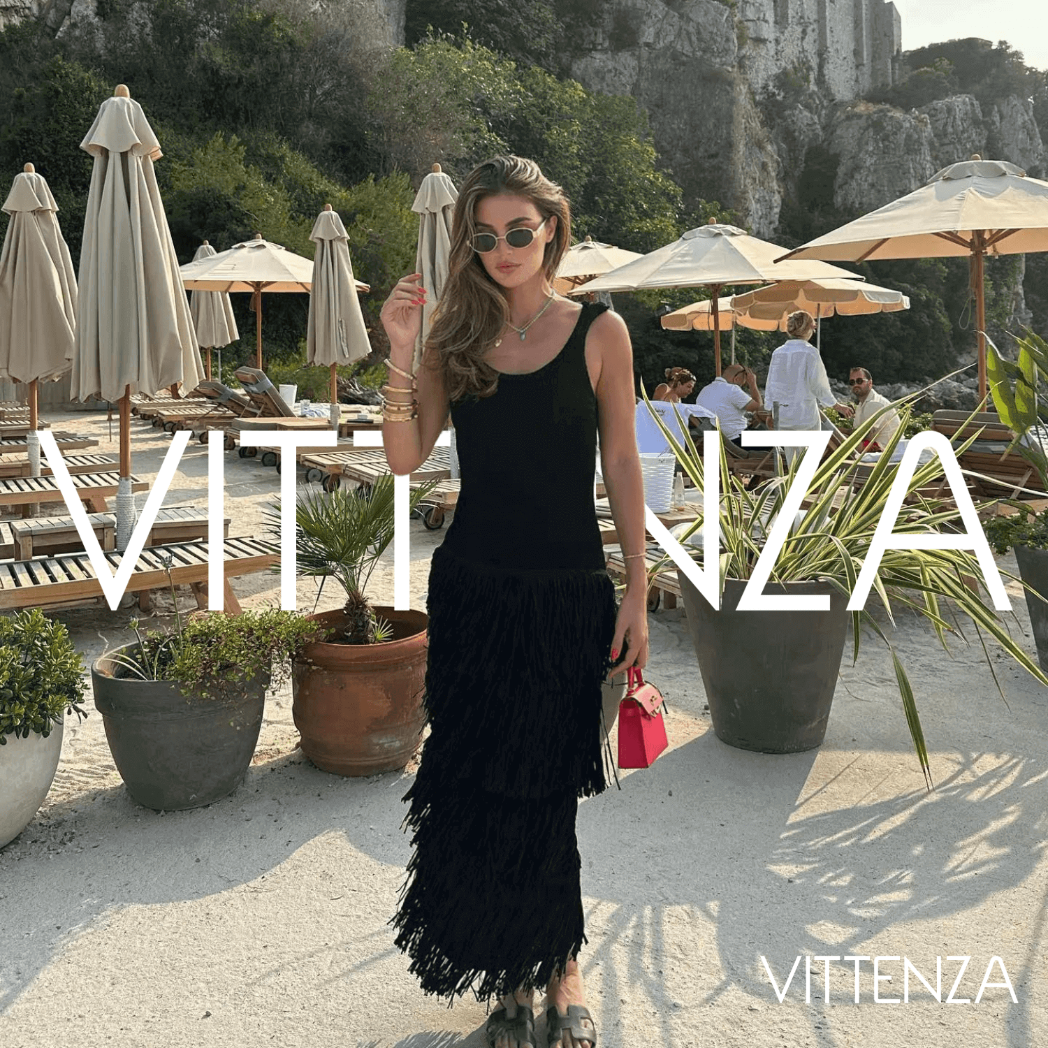 Front view of the black Bahamas dress worn by a model at a stylish beachside location, accessorized with a red handbag and statement sunglasses.