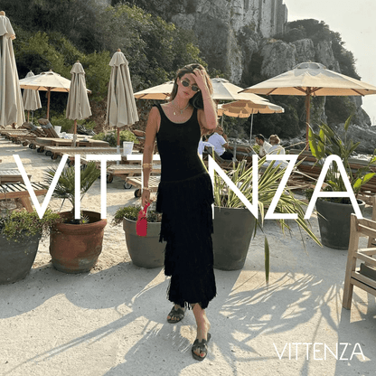 Model wearing the black Bahamas dress at a beachside venue during sunset, holding a red handbag and styled with sunglasses.