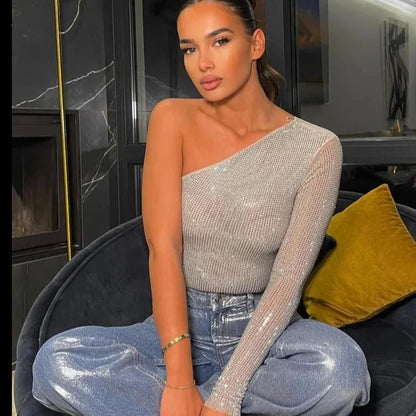 Woman seated in a modern living room, wearing a one-shoulder asymmetric sequin top paired with shimmering metallic jeans, showcasing a glamorous and contemporary style.